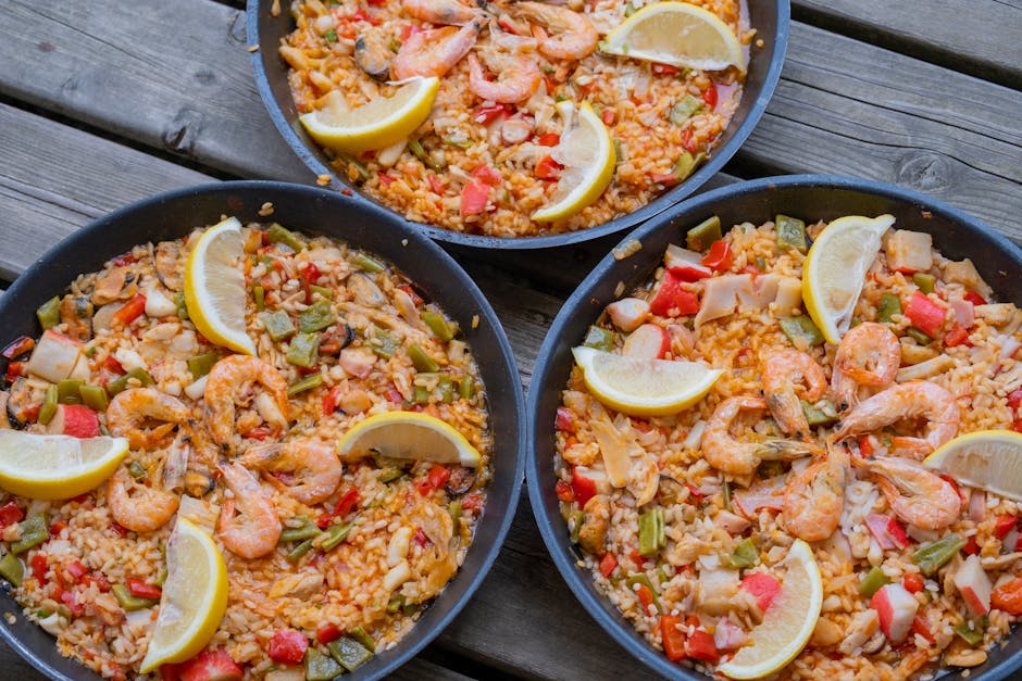Close-up of three pans of seafood paella with lemon slices on a wooden table.