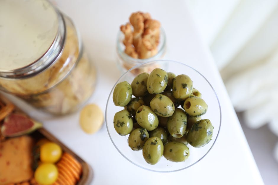 A detailed close-up of marinated green olives in a glass bowl with snacks in the background.
