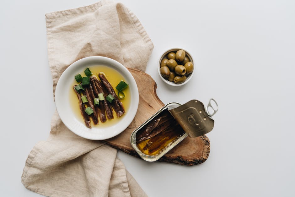 A flat lay of anchovies in oil, accompanied by green olives on a rustic wooden board.